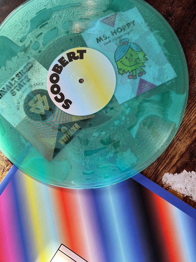 Close view of the handmade teal vinyl. Stickers read Unaltered State West Coast Pilsner, Wayfinder, and Ms. Hoppy. The center label reads SCOOBERT.