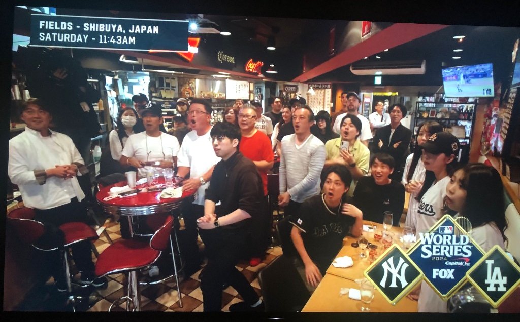 A photograph of a television. The screen shows a crowded bar in Shibuya, with a graphic reading Fields, Shibuya, Japan, Saturday, 11:43 a.m., and the FOX World Series 2024 logo for the Yankees and the Dodgers.